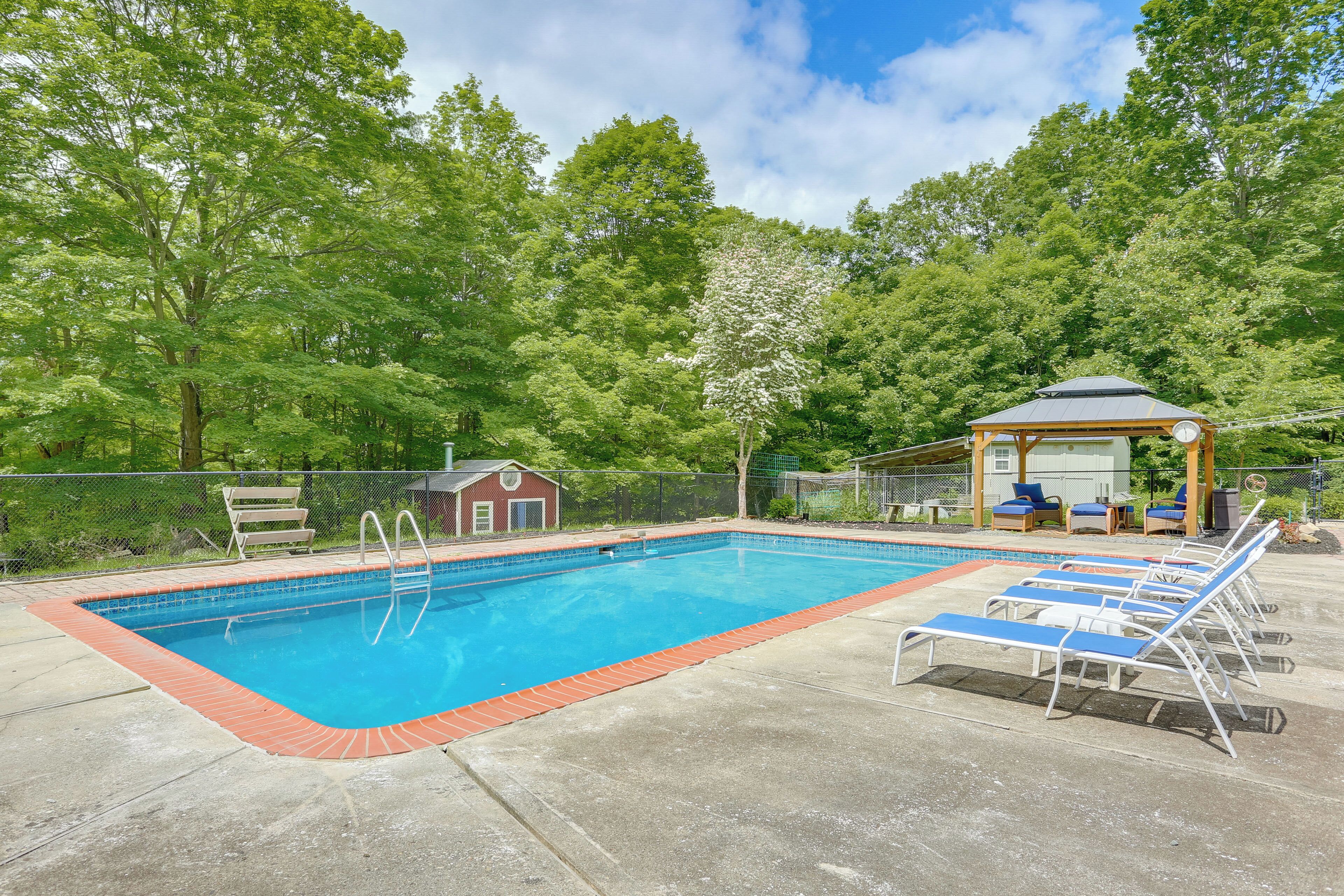 A rectangular swimming pool with a red border, surrounded by a concrete deck. There are lounge chairs and a gazebo with a roof.