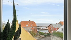 Vue sur une ville côtière depuis une fenêtre, avec un bâtiment jaune, une maison à toit rouge et une étendue d’eau.