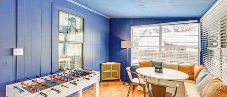 Ein Zimmer mit Kicker, einem Fenster mit Jalousien, einem runden Tisch mit Stühlen und einer Bank mit Kissen.