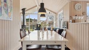 Un espace repas comprenant une longue table, des chaises, des luminaires suspendus, une horloge et une vue dégagée grâce à de grandes fenêtres.