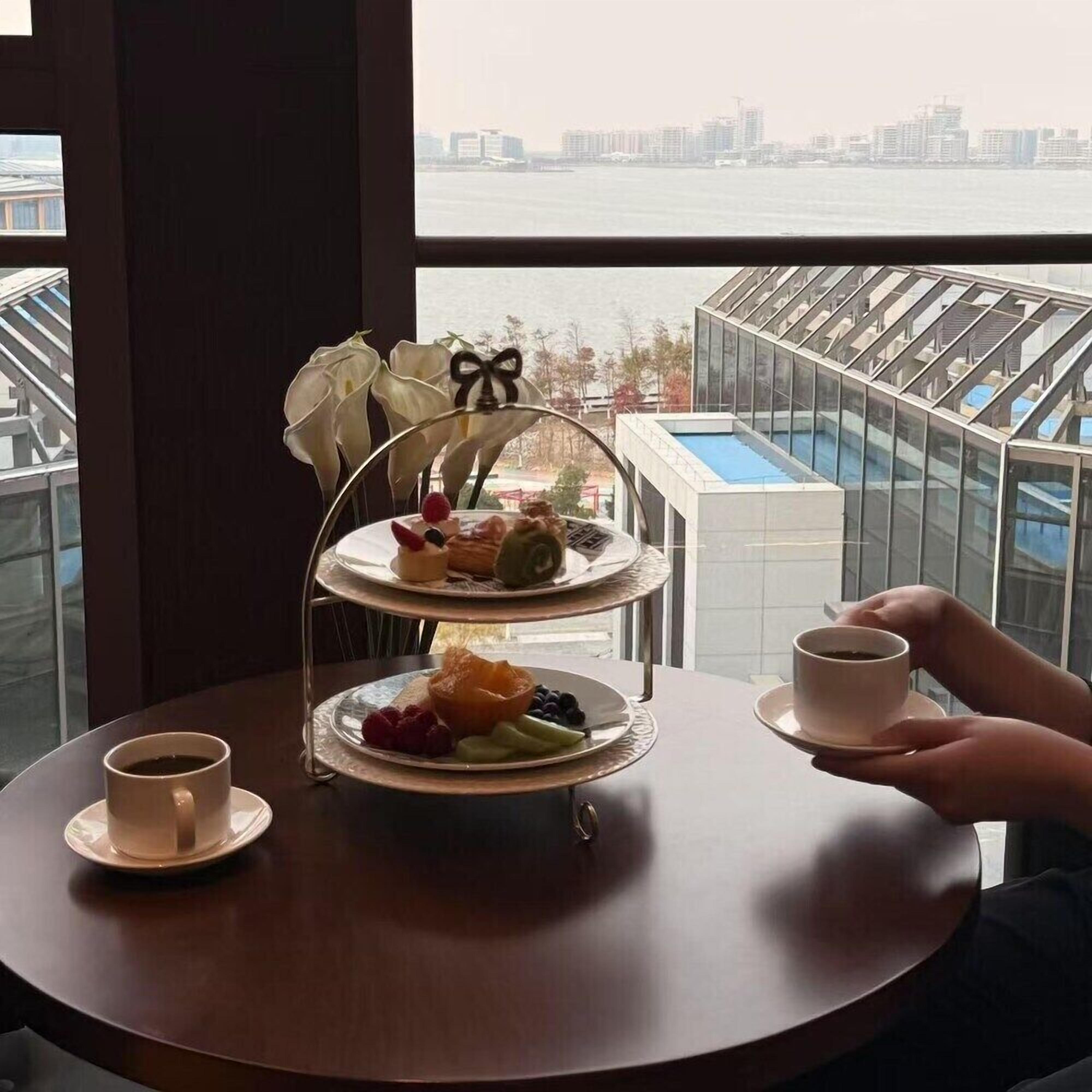Un présentoir à trois niveaux garni de diverses pâtisseries et de fruits, une tasse de café et une vue sur les toits d’une ville, le tout visible à travers une grande fenêtre.