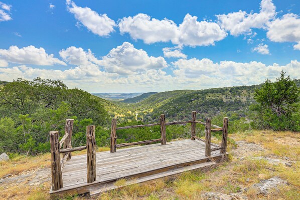 A wooden platform with a metal railing overlooking a valley with dense greenery and a clear sky. - Fire Pit, Views: Peaceful Texas Hill Country Cabin (Tarpley)