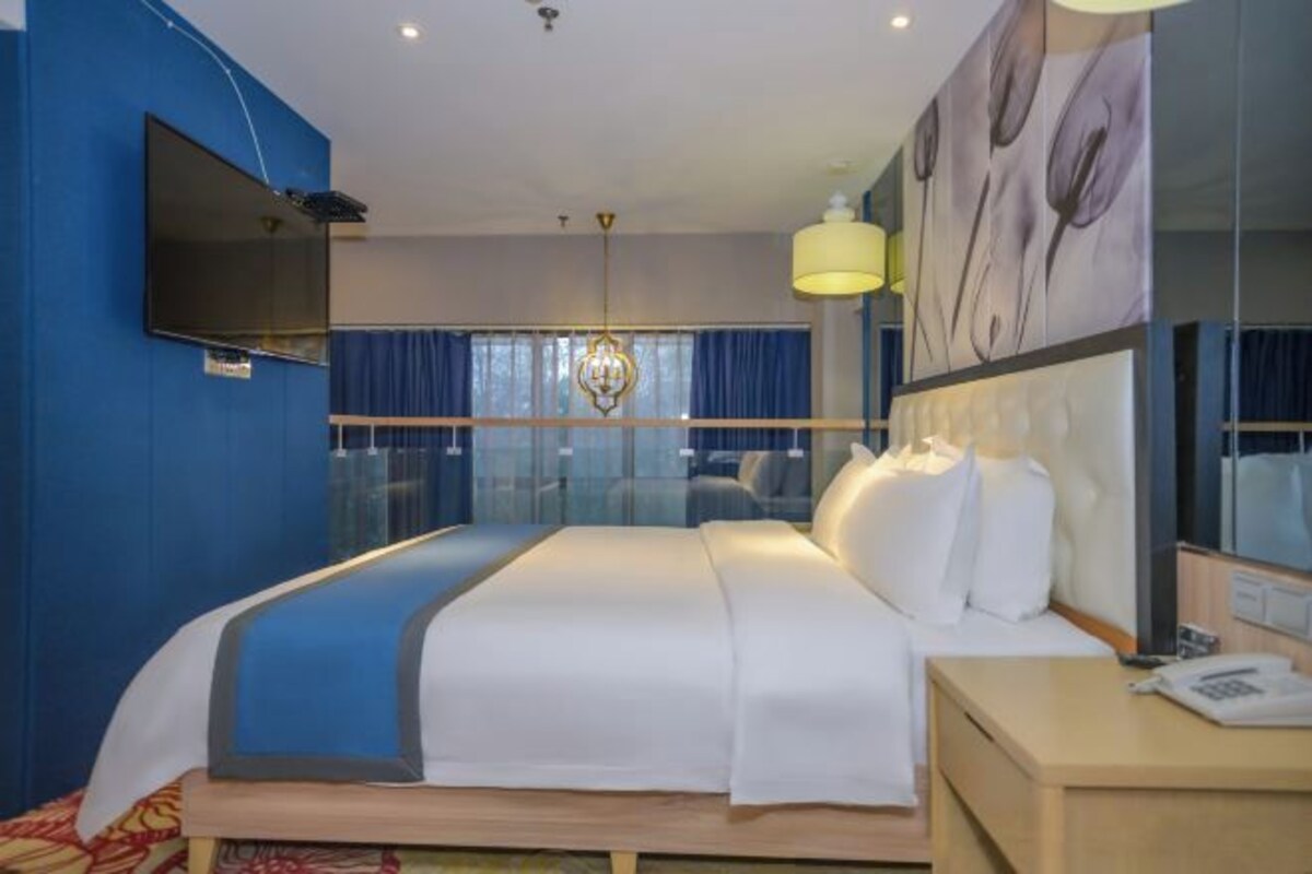 A hotel room with a large bed, a flat-screen TV mounted on the wall, a bedside table with a lamp, and a large window with curtains.
