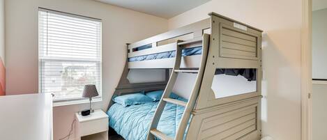 A bunk bed with a desk and lamp, a window with blinds, and a trash can.