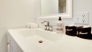 Un lavabo blanc dans une salle de bain, avec un miroir au-dessus, un flacon de savon liquide et deux serviettes noires sur lesquelles est inscrit le mot « Cosmétique ».