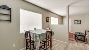 Un coin repas avec une table et des chaises, une fenêtre avec des stores, une porte et une petite table sur laquelle se trouve un plateau.