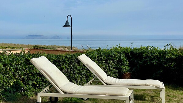 Two white lounge chairs with cushions, positioned on a grassy area with a view of the sea and distant landforms.