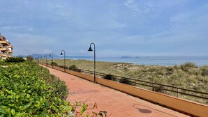 Un sendero pavimentado con farolas, bordeado de follaje y vista al mar y colinas distantes.