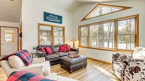 A spacious living room with a large window, a leather sofa, a white armchair, and a wooden ceiling.