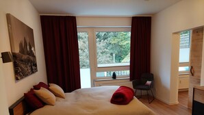 A bedroom with a bed, red curtains, a window with a view of trees, a chair, and a desk.