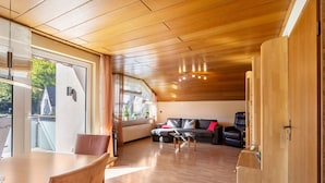 A modern living room with a wooden ceiling, a sofa, a dining table, and a view through a large window. - 'Urban' Apartment with Mountain View, Balcony & Wi-Fi – Tourist Use Only (Bestwig)