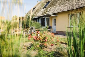 A thatched-roof building with a garden, outdoor seating, and a door with an anchor symbol.