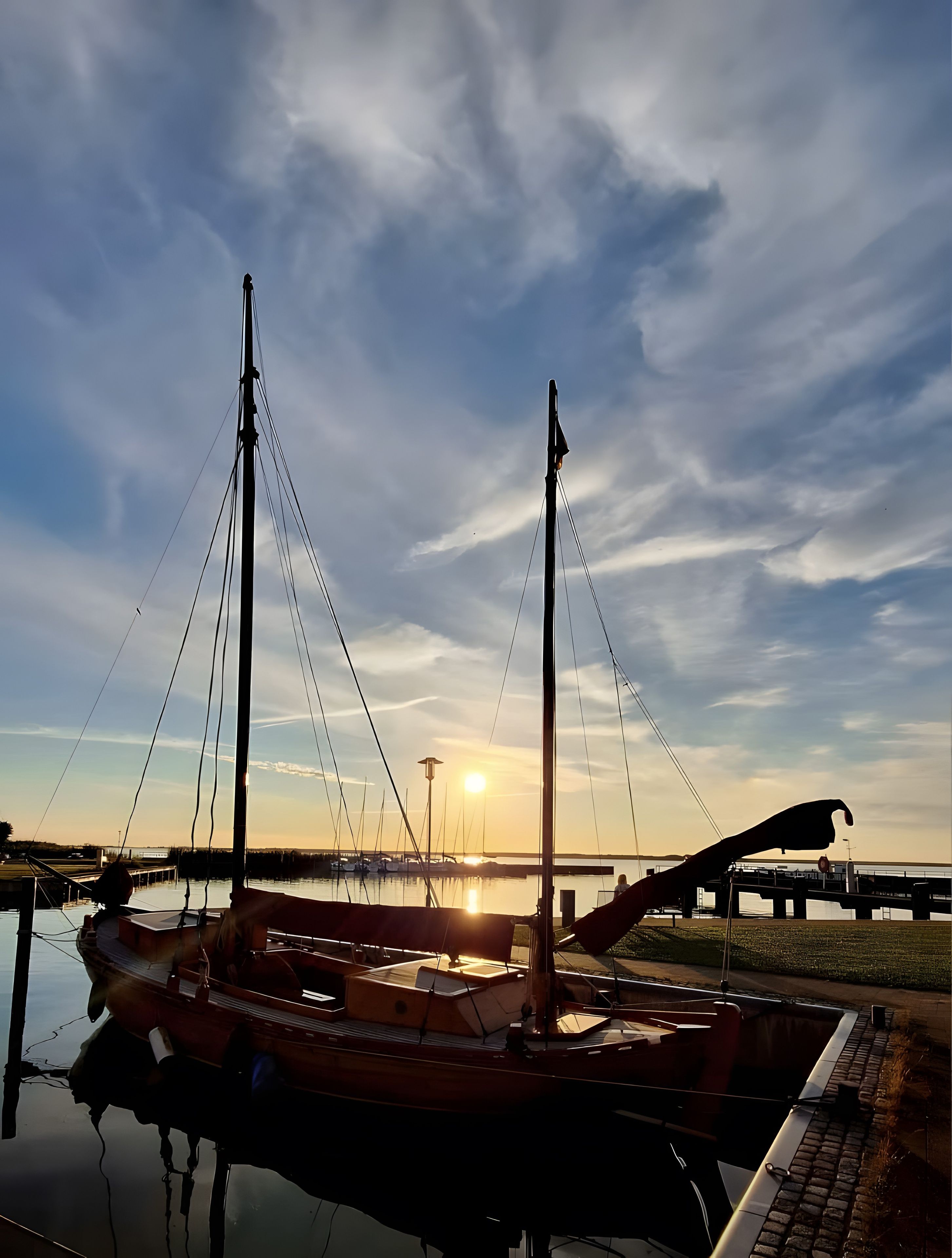 Ein an einem Steg festgemachtes Boot mit Masten und Tauwerk, ruhiges Gewässer und ein Sonnenuntergang im Hintergrund.