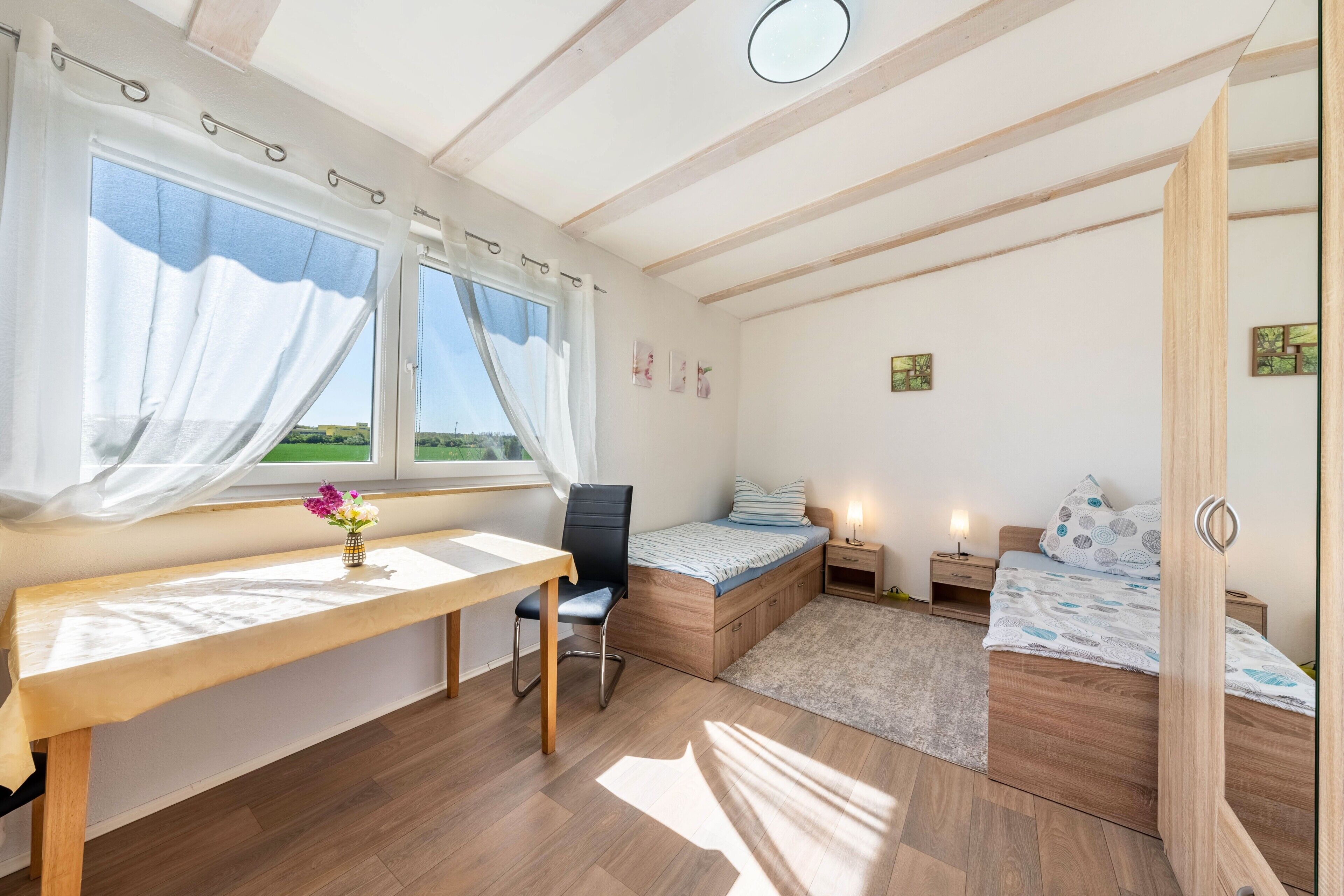 A bedroom with a wooden desk, a chair, a bed, a bedside table, and a window with curtains.