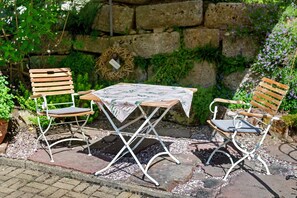 Un patio jardín con dos sillas de madera y una mesa, rodeado de follaje y un muro de piedra.