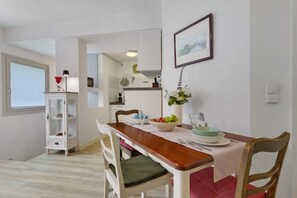 Un comedor con mesa de madera, sillas blancas, un jarrón con flores y una cocina al fondo.