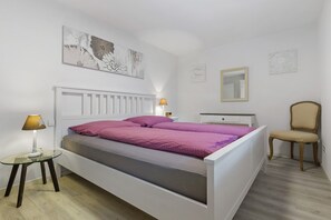 Una cama blanca con cabecera, una mesita de noche con lámpara, una silla y cuadros en las paredes.