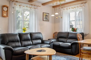 Ett vardagsrum med två svarta lädersoffor, ett träbord och ett matbord med stolar. Det finns två fönster med gardiner, en klocka på väggen och en inramad tavla ovanför soffan.
