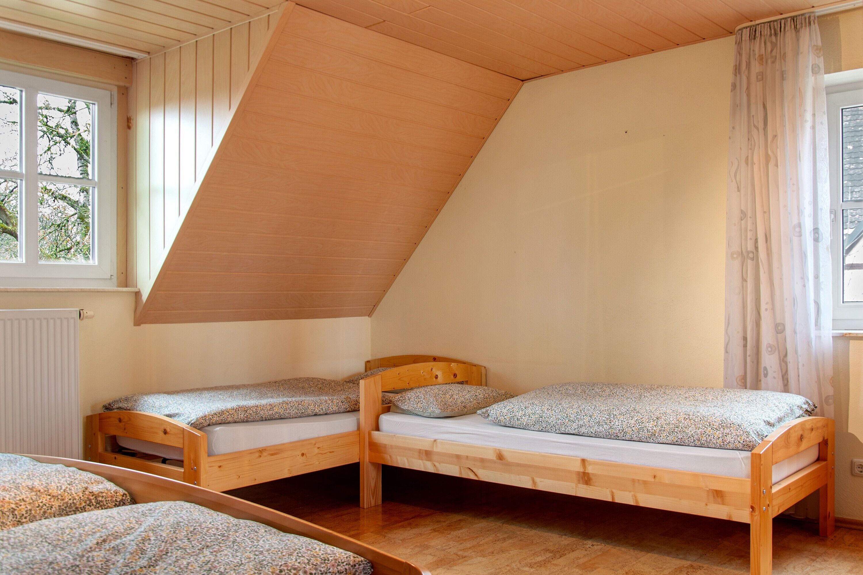Una habitación con dos camas literas de madera, techo inclinado, ventana con vistas a árboles y un radiador.