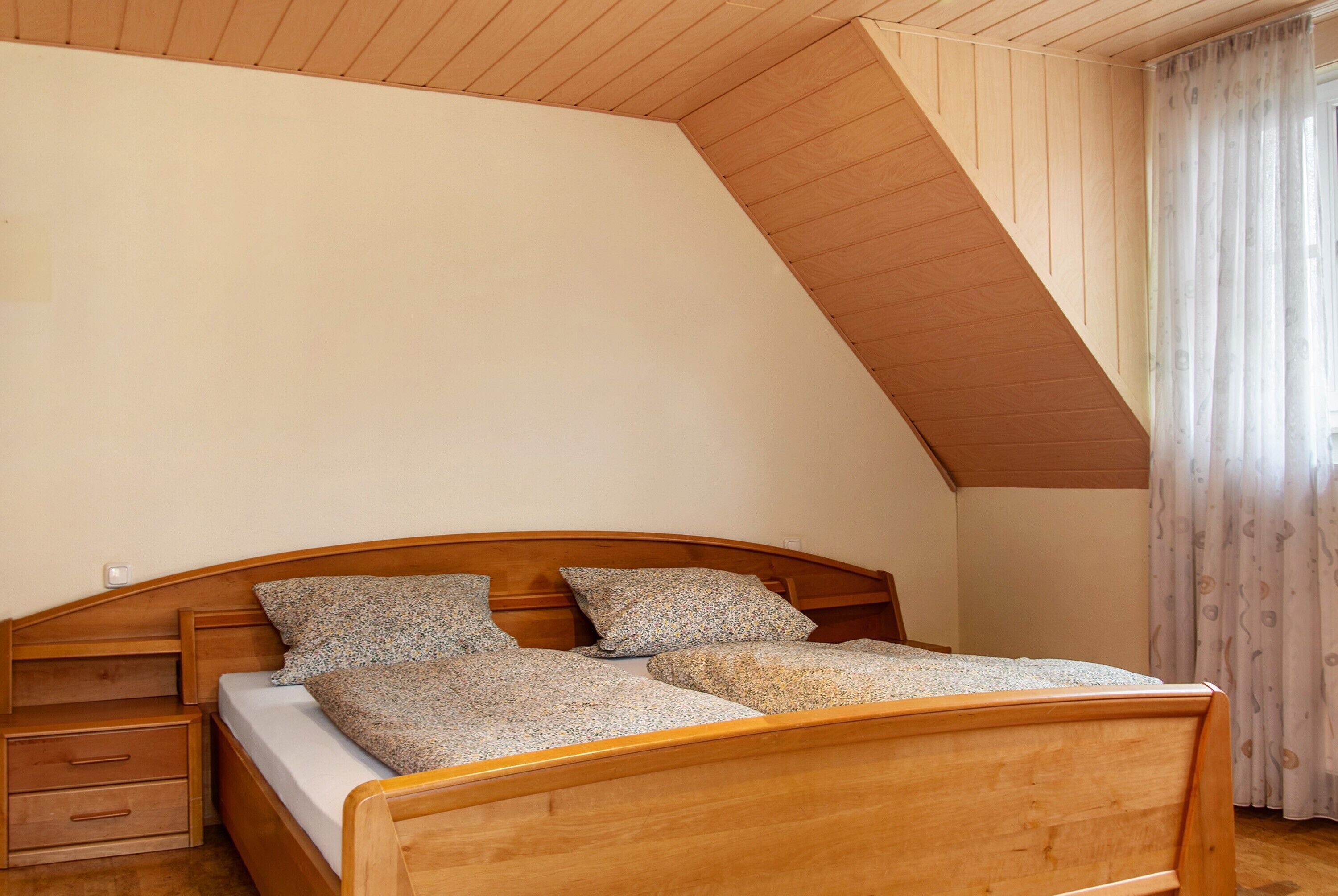 Ein Schlafzimmer mit einem hölzernen Bett, zwei Kissen, einem Nachttisch und einem Fenster mit Vorhängen.