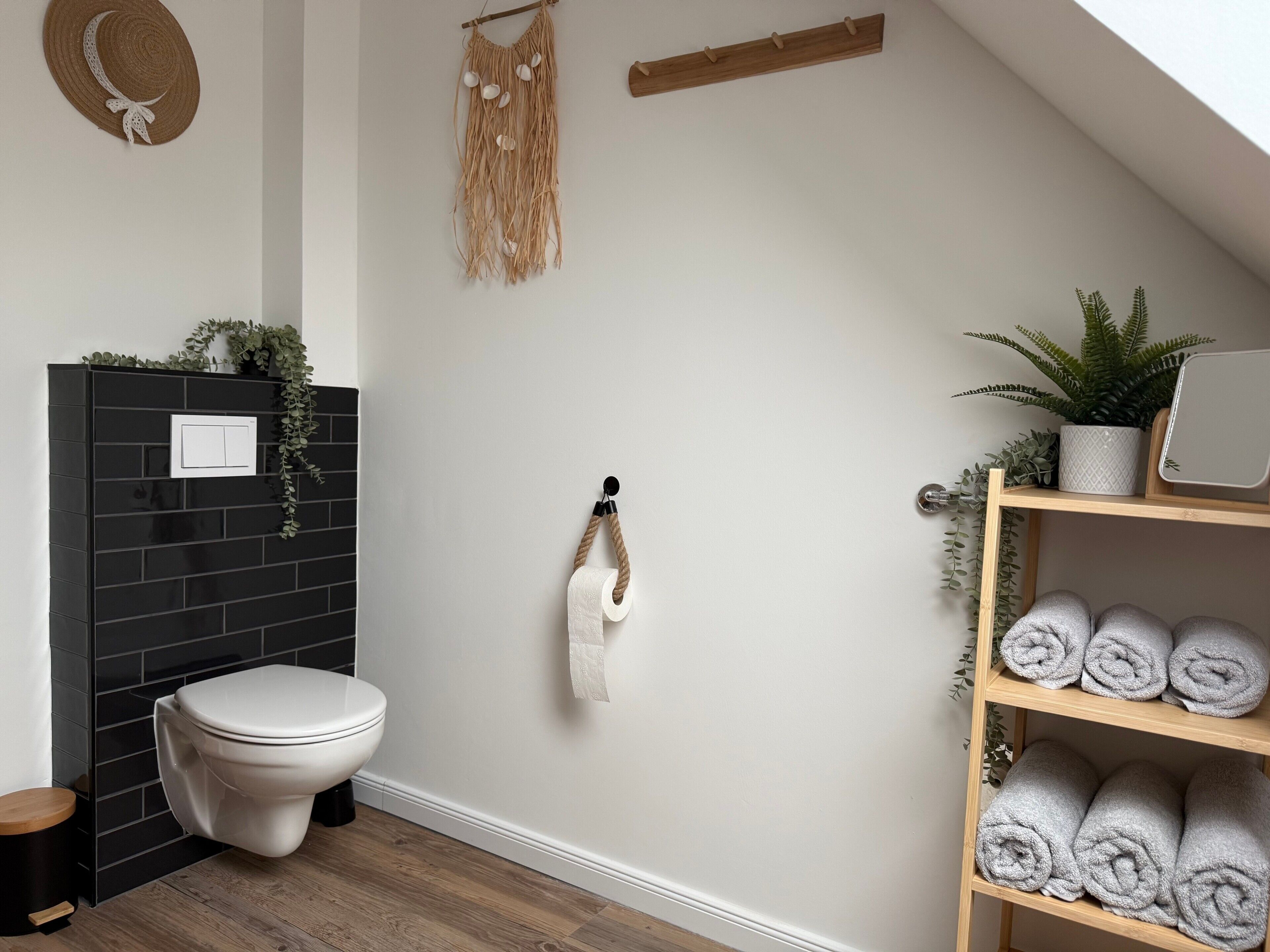Ein Badezimmer mit Toilette, Handtuchhalter, Ablagefläche mit aufgerollten Handtüchern und einer Wandpflanze.