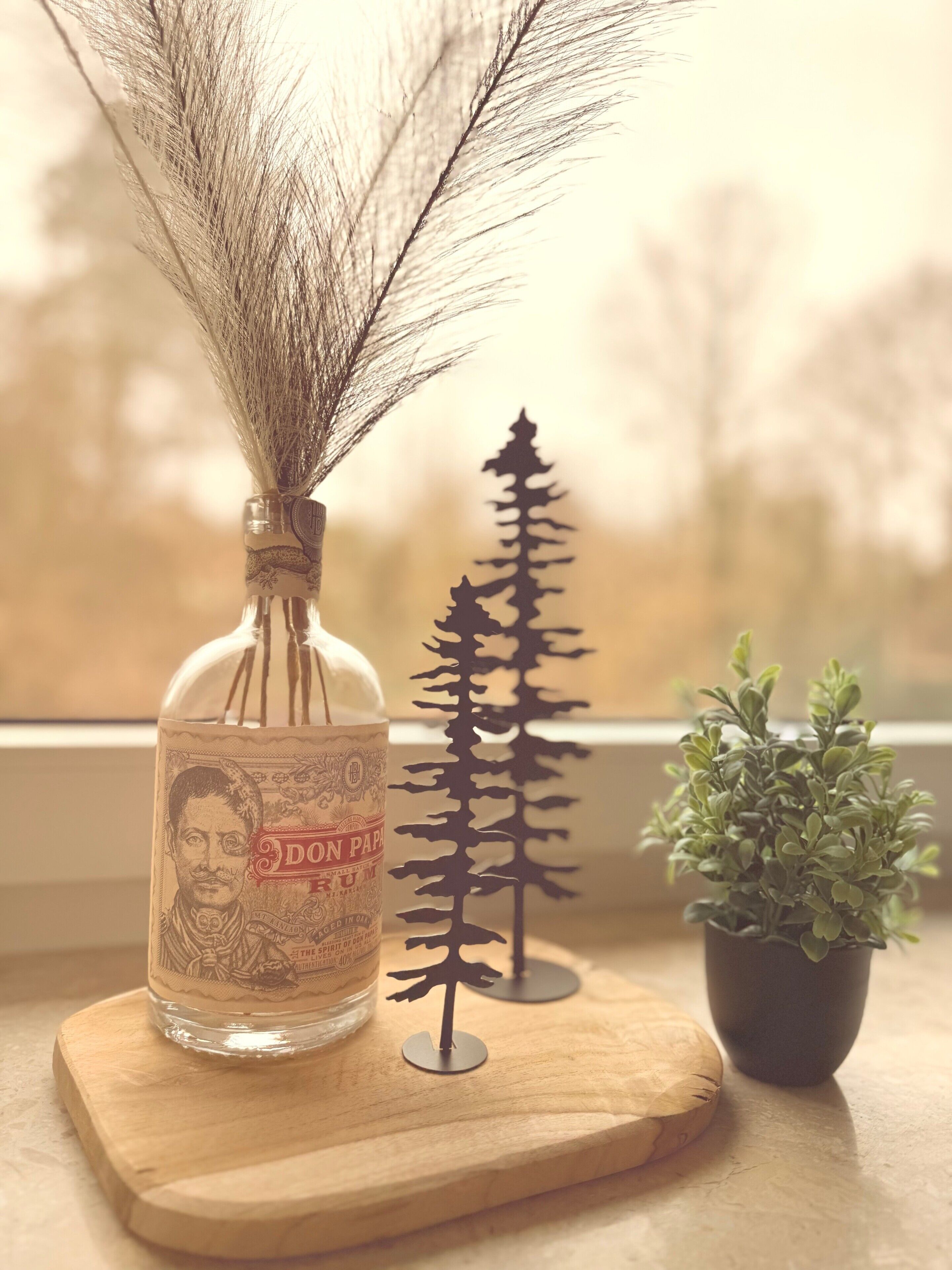 Eine Flasche mit Etikett, eine kleine Baumdekoration und ein Topfpflanze auf einer Holzplanke.