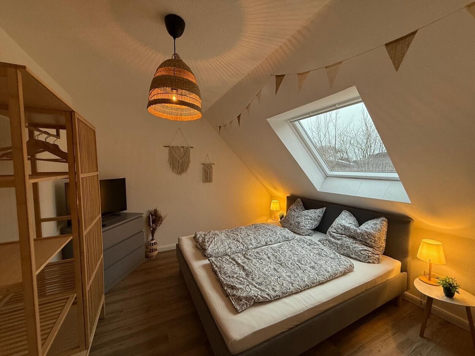 Een slaapkamer met een schuine wand, een bed met beddengoed met een patroon, een hangende lamp, een kledingkast, een klein tafeltje met een lamp en een raam met uitzicht.
