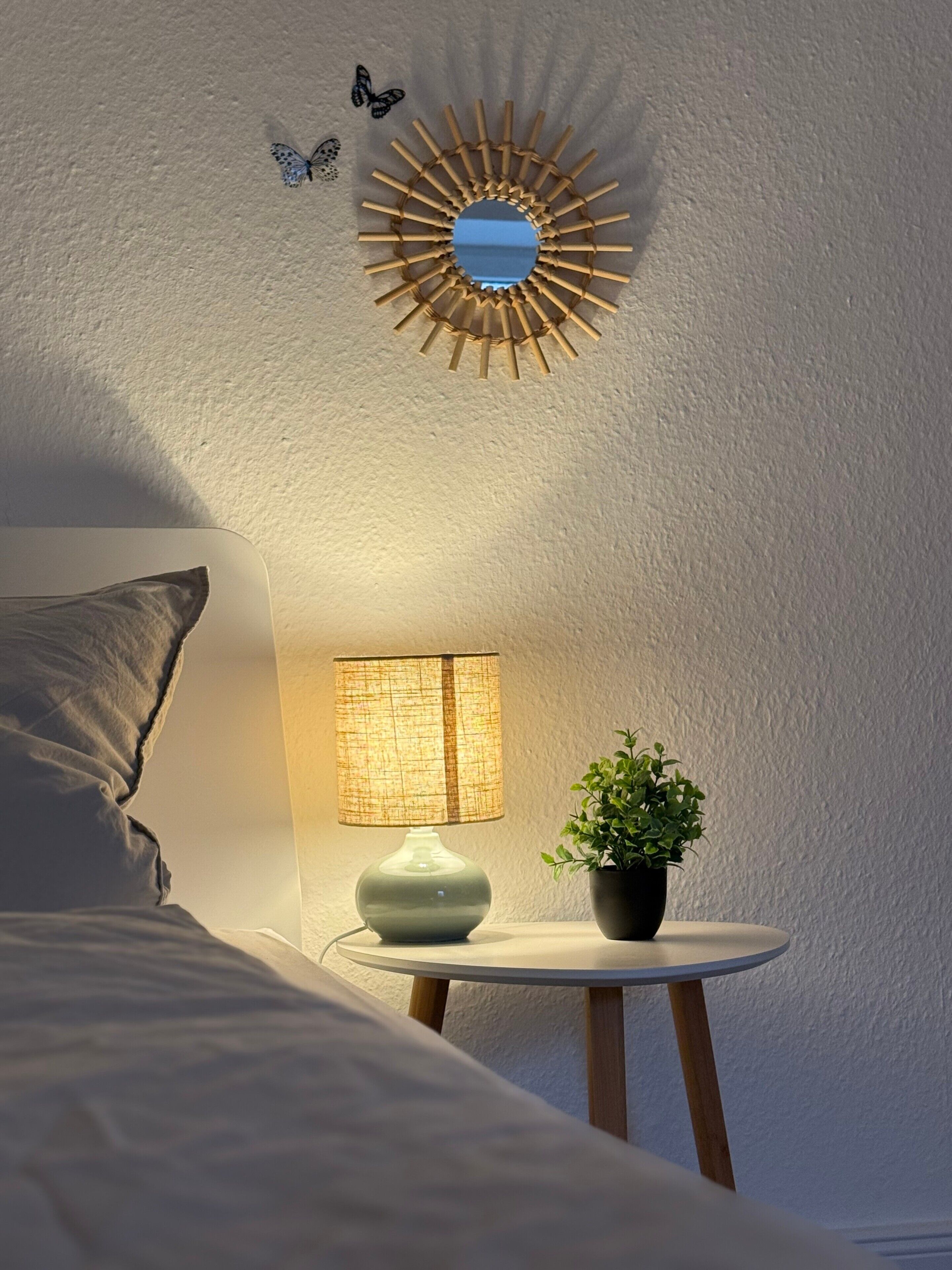 A bedroom with a bed, a bedside lamp, a small table with a plant, and a wall-mounted mirror.