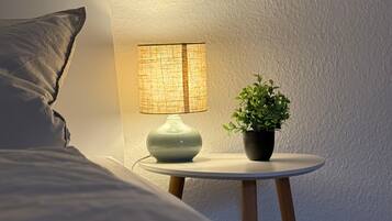 A bedroom with a bed, a bedside lamp, a small table with a plant, and a wall-mounted mirror.