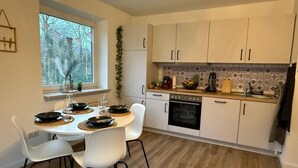 A modern kitchen with white cabinets, a dining table with chairs, an oven, and a window with a view of trees. - Apartment 'Hygge' with Private Terrace, Private Garden and Wi-Fi (Hennstedt)