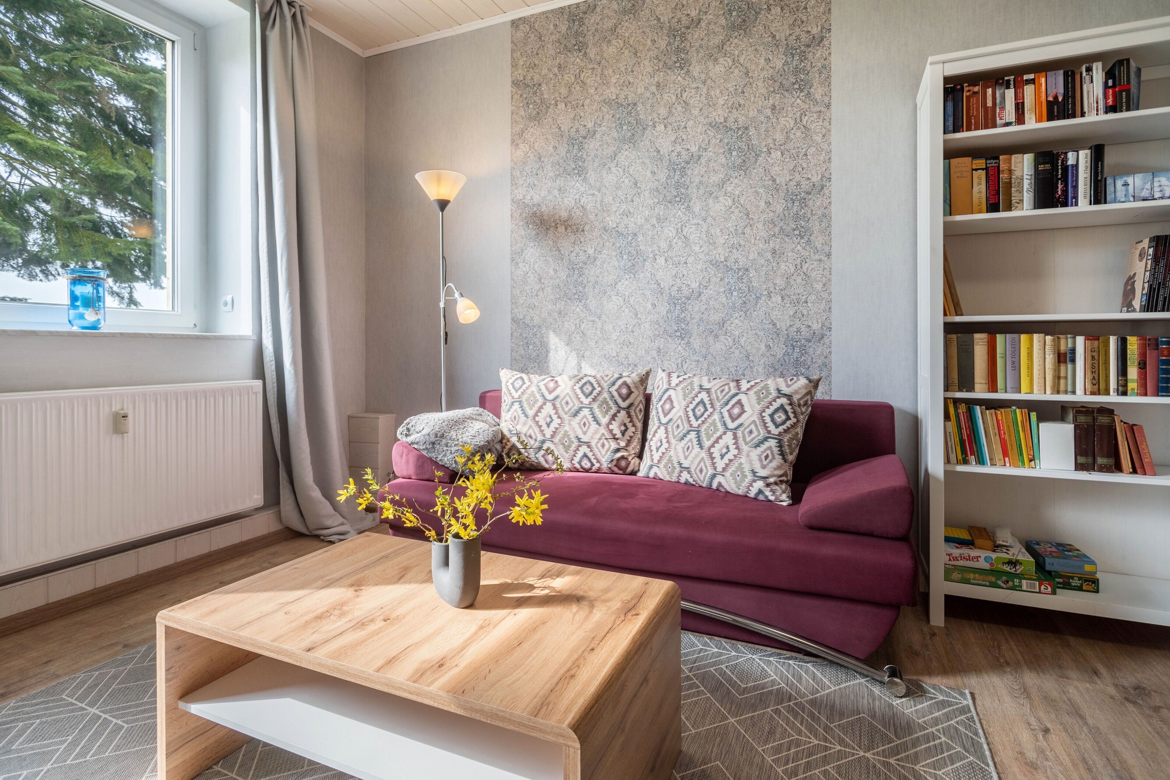 Een woonkamer met een houten salontafel, een rode bank met gekleurde kussens, een vloerlamp en een boekenplank vol boeken.