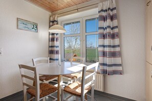 Uma área de refeições com uma mesa e cadeiras brancas, uma luminária pendente e uma janela com cortinas.
