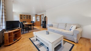 A living room with a white sofa, a TV on a wooden stand, a dining area with a table and chairs, and a kitchen area with cabinets and a stove.