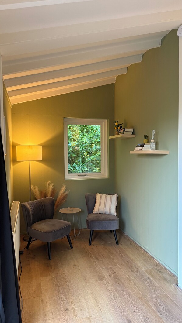 A cozy room with a wooden floor, two grey armchairs, a small round table, a floor lamp, a window with a view of greenery, and a shelf with decorative items.