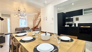 Un espace de salle à manger moderne avec une table en bois dressée pour quatre personnes, un escalier et une cuisine en arrière-plan.