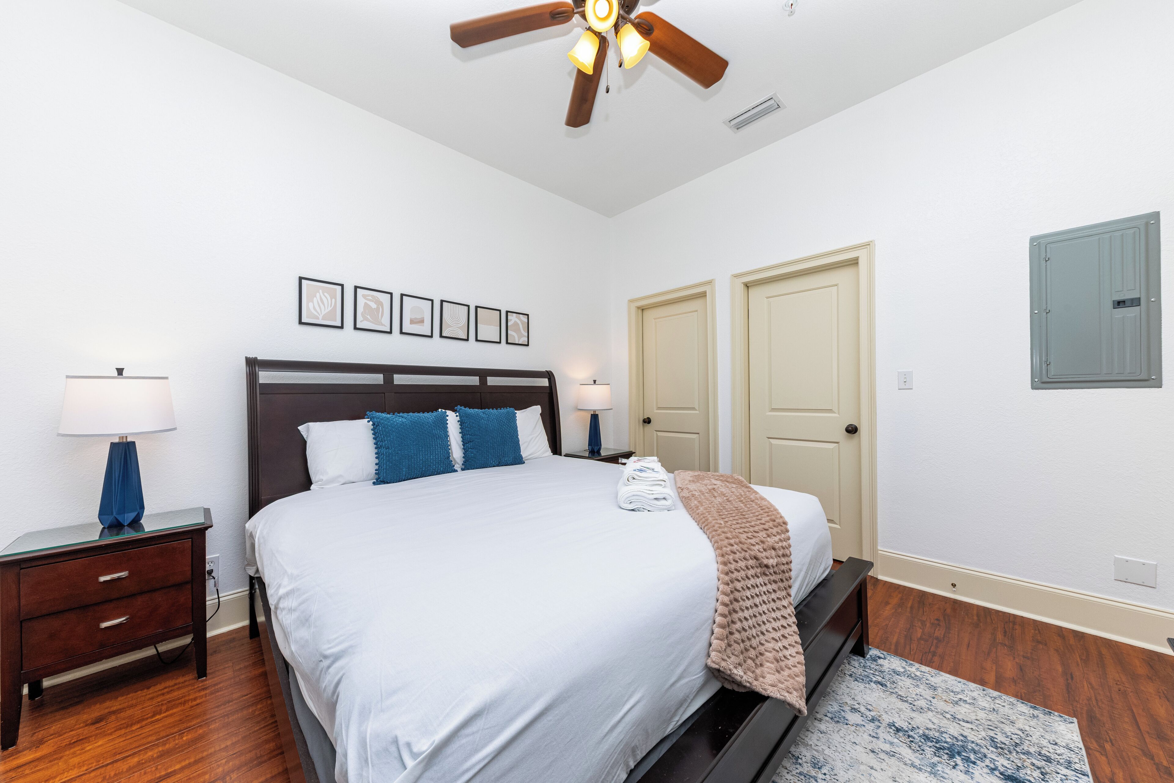 Un dormitorio con cama, mesitas de noche, ventilador de techo, cuadros enmarcados y puertas.