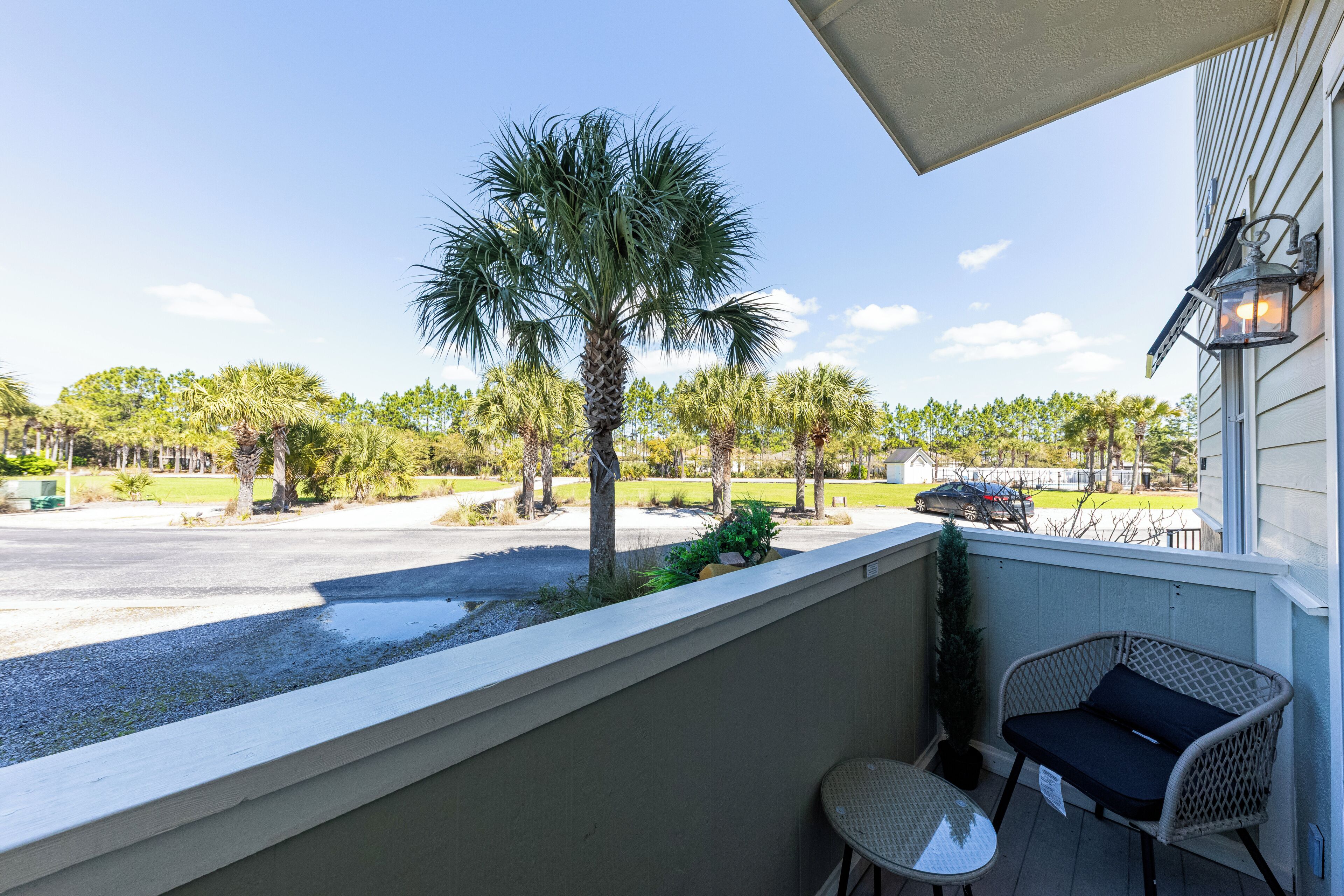 A balcony with a wicker chair and table, overlooking a street with palm trees and a parked car.