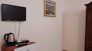 Une petite pièce avec un réfrigérateur blanc, un téléviseur fixé au mur, un tableau encadré et un tapis à motifs sur le sol.