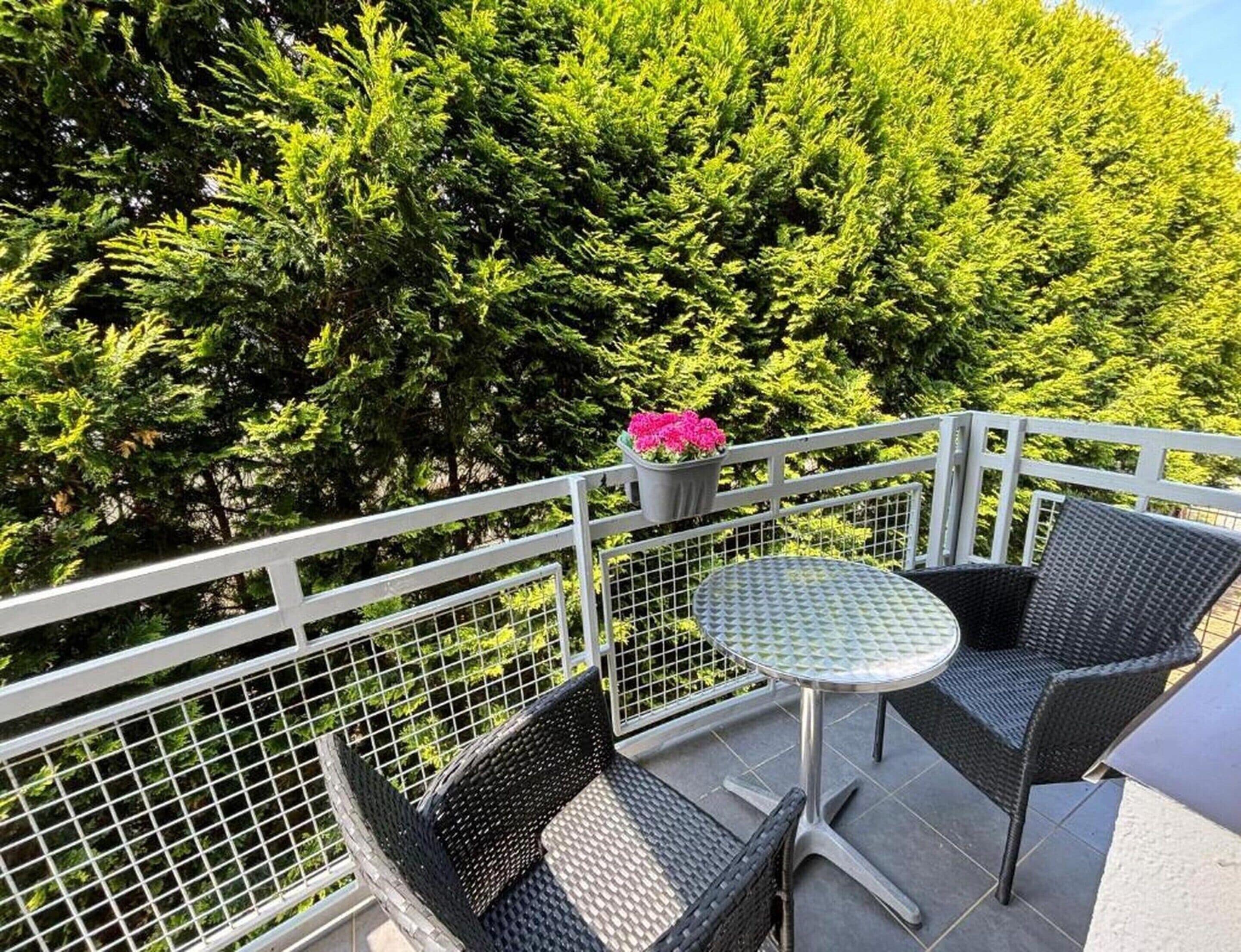 A balcony with a metal railing, a small round table, two wicker chairs, and a potted plant with pink flowers.