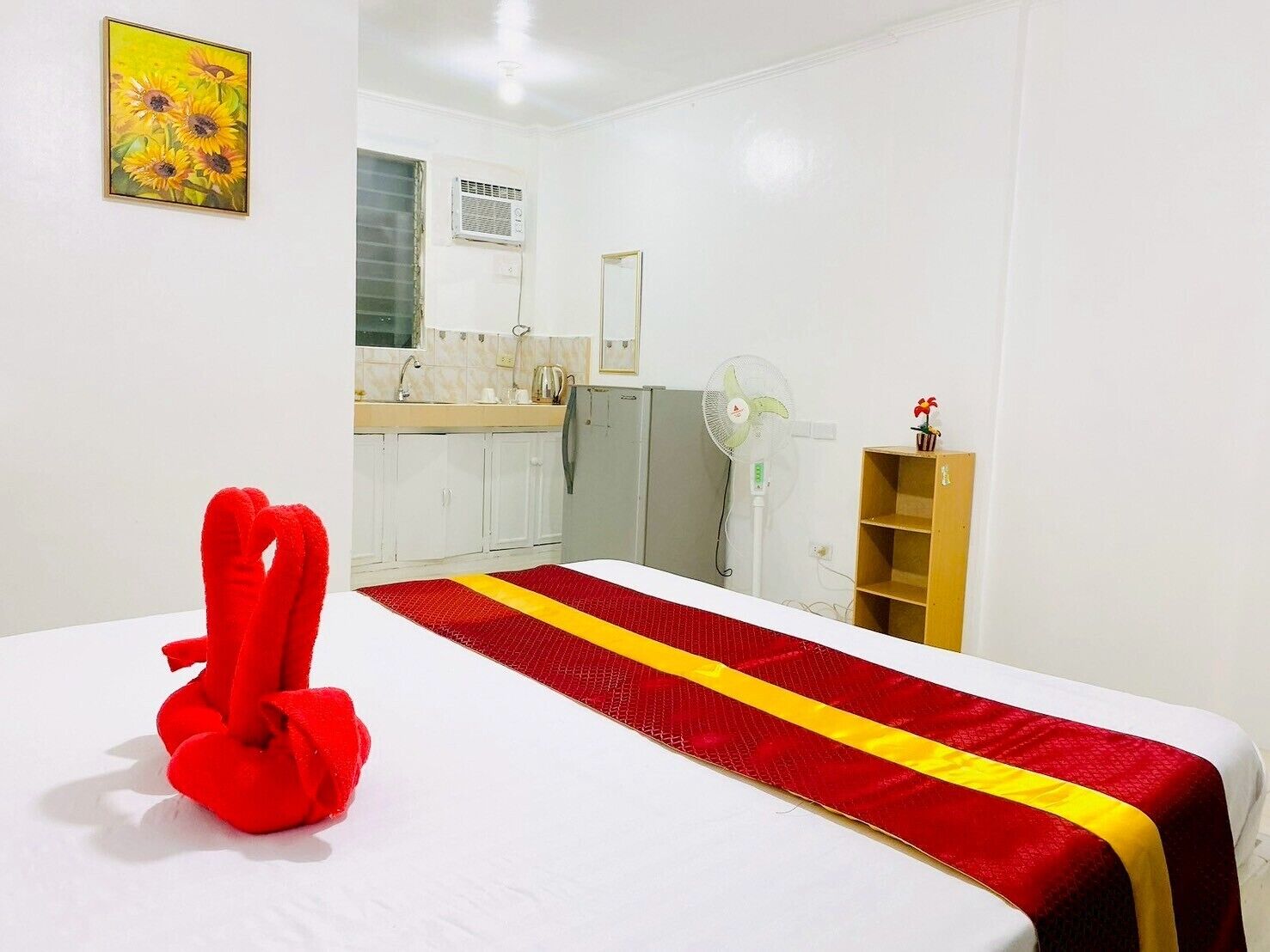 A hotel room with a bed, a red towel, a kitchen area, and a small shelf with a plant.