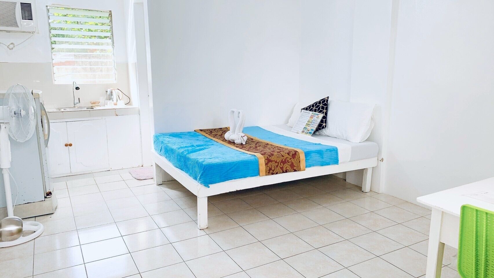 A clean, white room with a bed, a small table, and a fan.