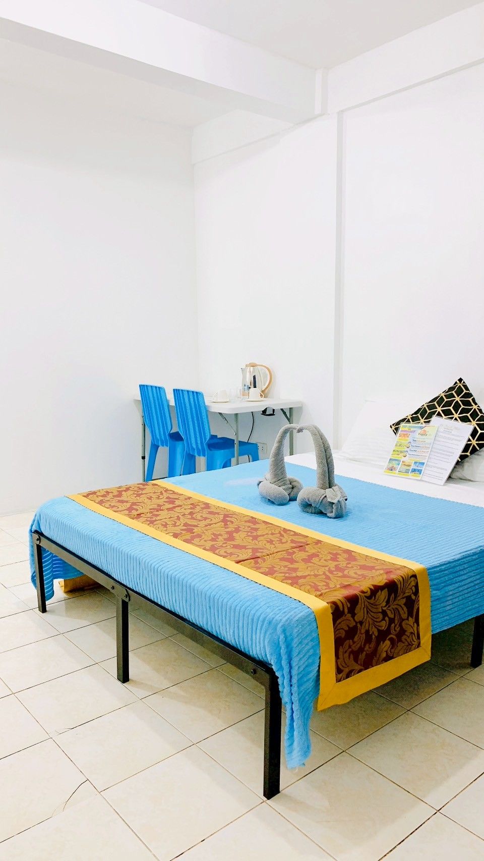 A single bed with a blue and red patterned bedspread, a small desk with a chair, a kettle, and a mirror.