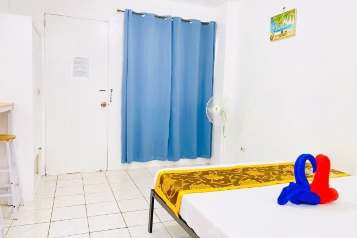 A room with a bed, a desk, a chair, a blue curtain, and a fan.