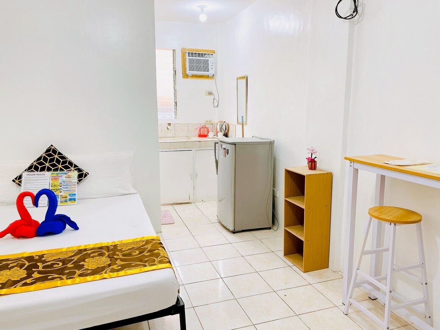 A small, clean room with a bed, a desk, a stool, a refrigerator, and a bathroom visible in the background.