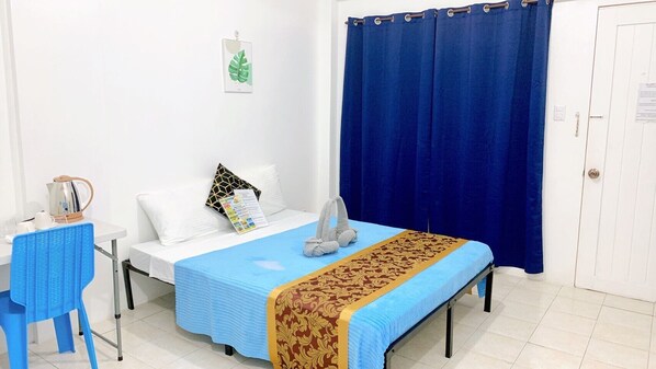 A hotel room with a bed, a table with a kettle, a chair, and a blue curtain.