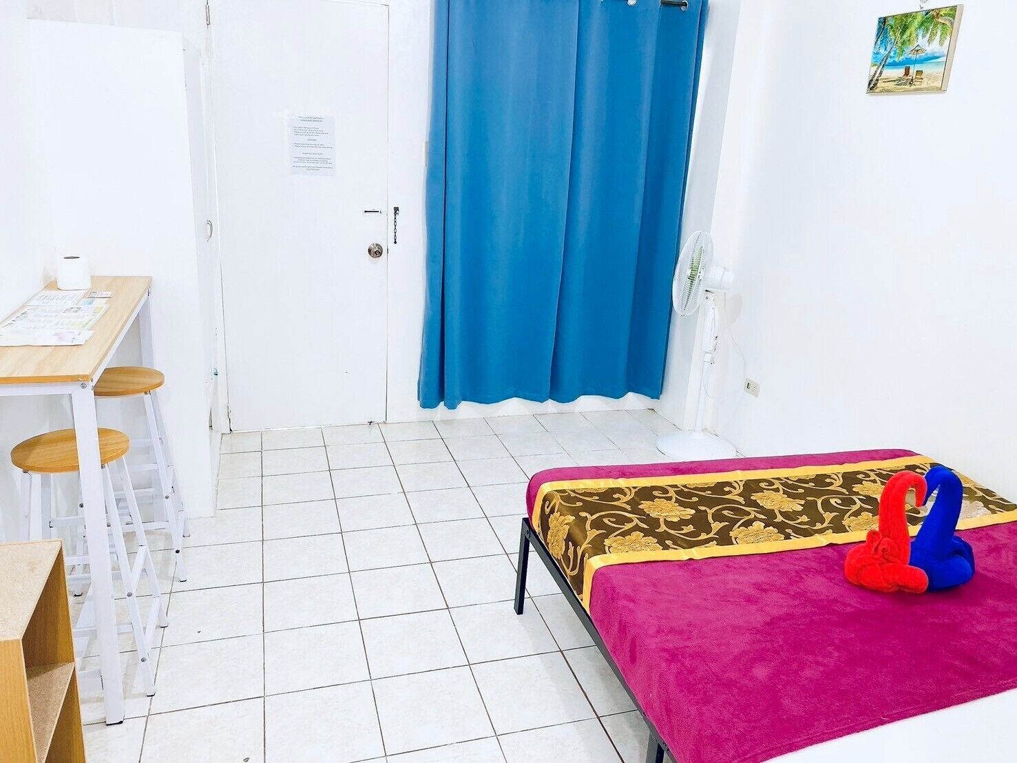 A small, clean room with a bed, a desk with chairs, a blue curtain, and a fan.