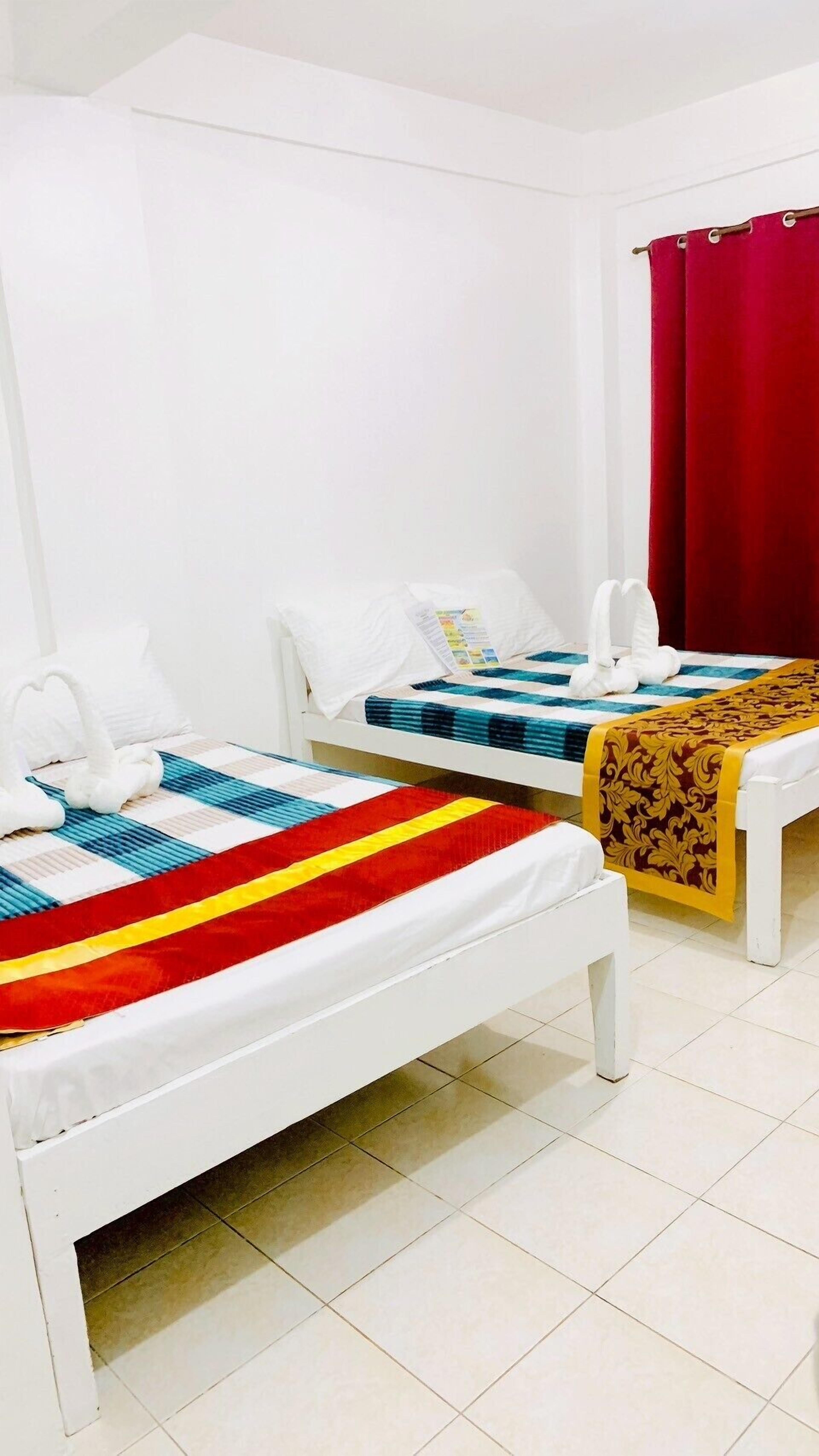 Dos camas individuales con estructura blanca y ropa de cama colorida, una cortina roja y una pared blanca.