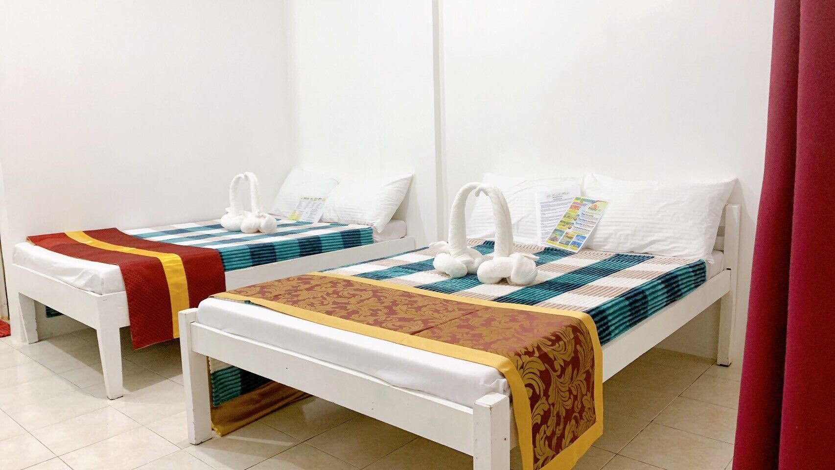 Dos camas individuales con estructura blanca y ropa de cama de colores, una cortina roja y una pared blanca.