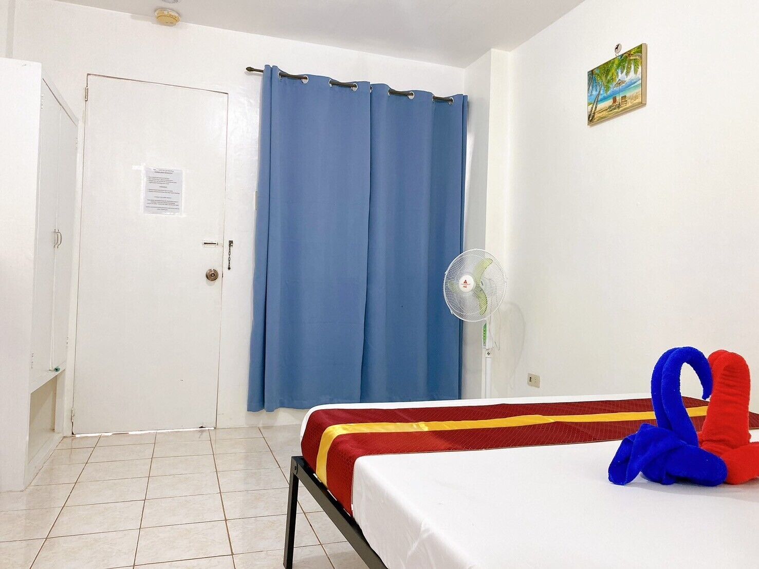 A room with a bed, a door, a blue curtain, a fan, and a small painting on the wall.
