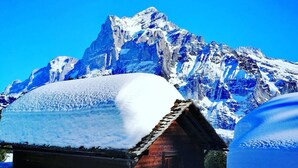 Una cabaña de madera con techo nevado, rodeada de nieve y montañas bajo un cielo azul despejado.